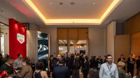 Busy trade show lobby with professionals mingling near brand booths; red TAG Heuer display on the left.