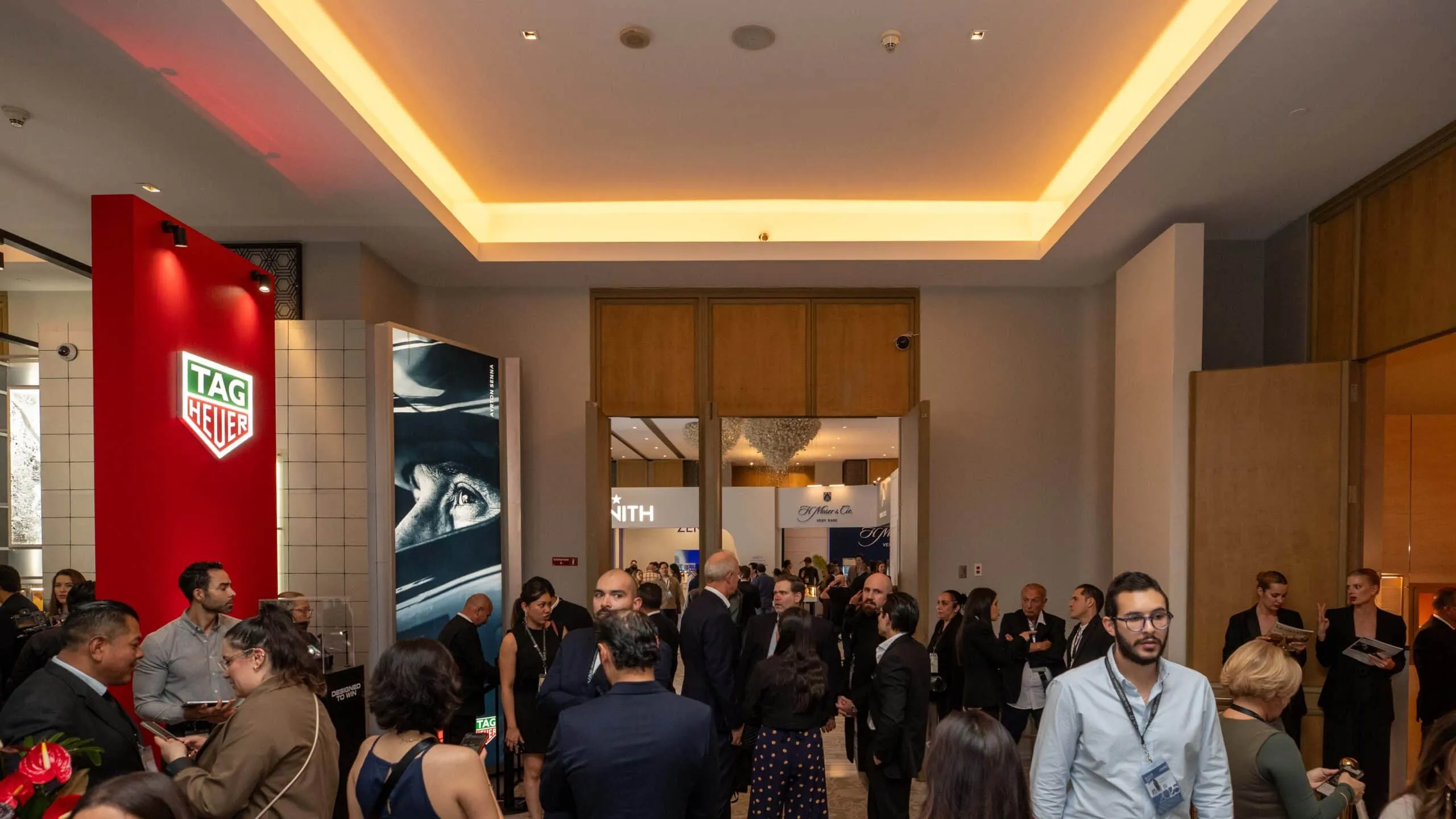 Busy trade show lobby with professionals mingling near brand booths; red TAG Heuer display on the left.