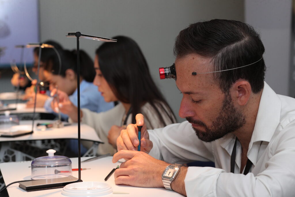 A10I0554 - Petróleo y Energía Scientists at a lab bench using precision tools with head-mounted magnifiers; focused on tasks in a classroom lab setting.