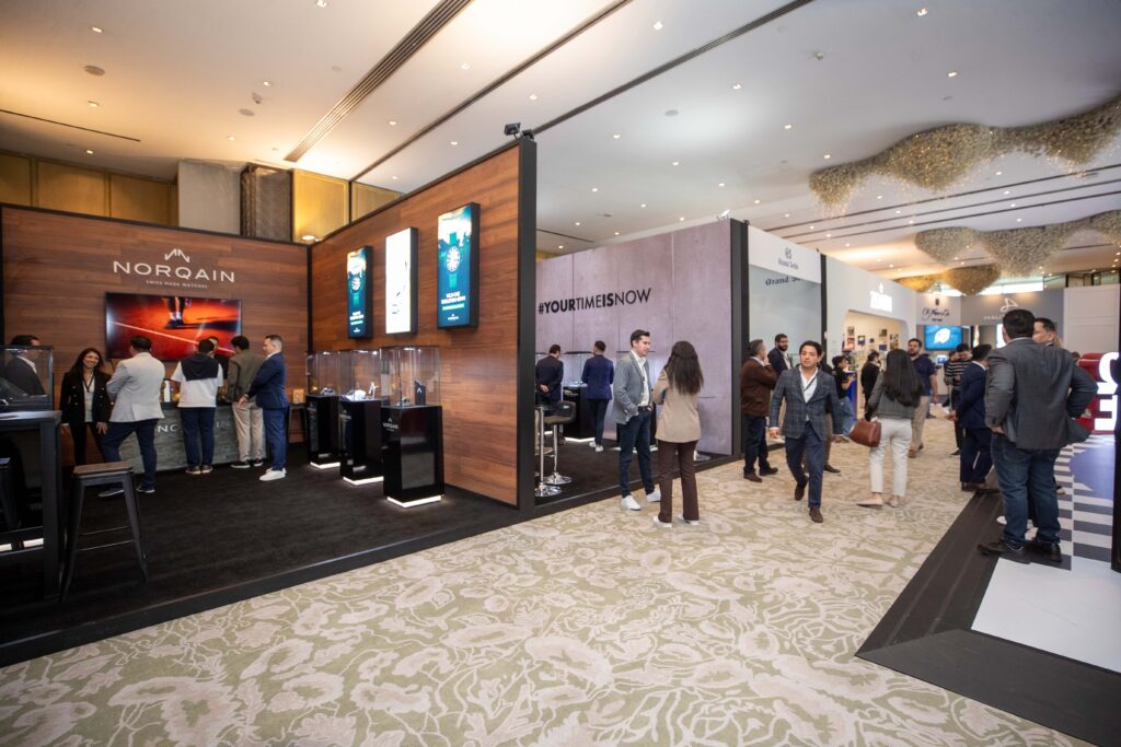 A10I0644 - Petróleo y Energía Busy exhibition hall with a Norqain booth featuring wood paneling, digital displays, and attendees talking and browsing displays.