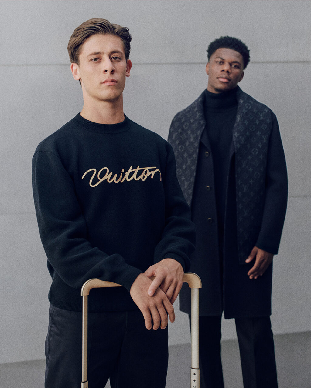 Arda_Guler_Aurelien_Tchouameni_Louis_Vuitton_Real_Madrid - Petróleo y Energía Two male models pose in a studio; foreground model wears a black sweatshirt with gold script logo, holding a suitcase handle. In the background, another model wears a dark coat with a monogram pattern and a black turtleneck.