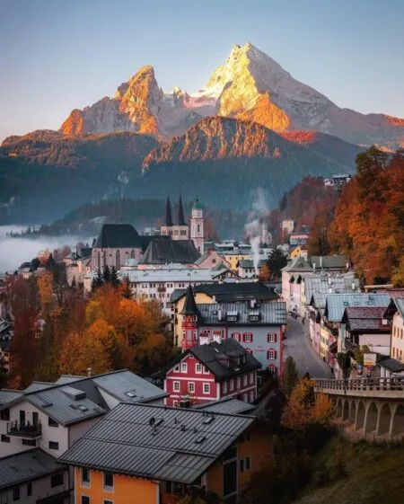 Autumnal alpine village with colorful houses and a church, set against snow-capped mountains at sunrise/early morning light.