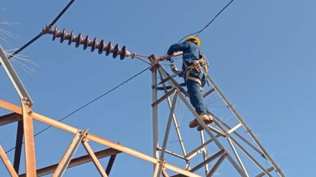 Worker in a yellow hard hat and safety harness atop a metal power-­line tower, performing maintenance on an overhead electrical conductor against a clear blue sky.