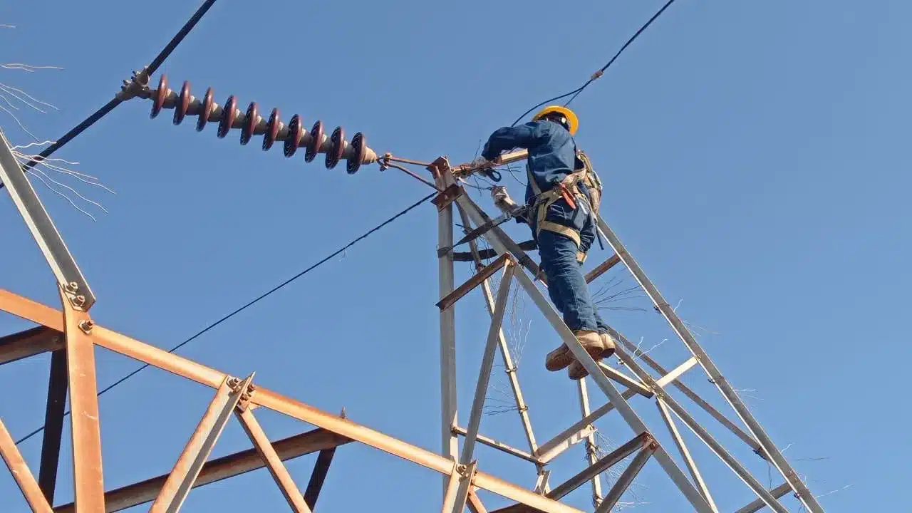 Worker in a yellow hard hat and safety harness atop a metal power-­line tower, performing maintenance on an overhead electrical conductor against a clear blue sky.