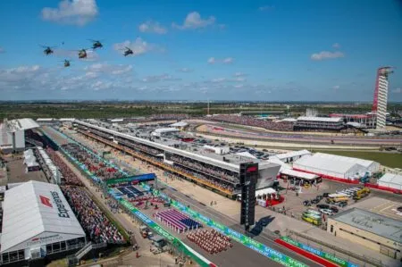 Aerial view of a crowded racetrack event with grandstands, pit buildings, and several helicopters flying overhead.