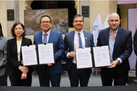 Four professionals in suits hold signed documents during a formal signing ceremony, with a map display and flags in the background.