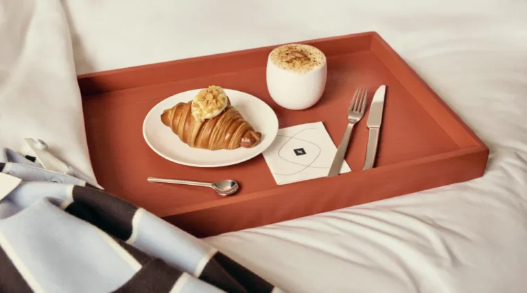 A white plate with a croissant topped by a dollop of cream sits on a red bedside tray, beside a white mug with frothy coffee on the tray.