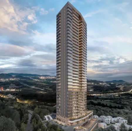 Tall, modern glass high-rise apartment tower rises above a wooded landscape against a blue sky at dusk, with many balconies visible on every floor.