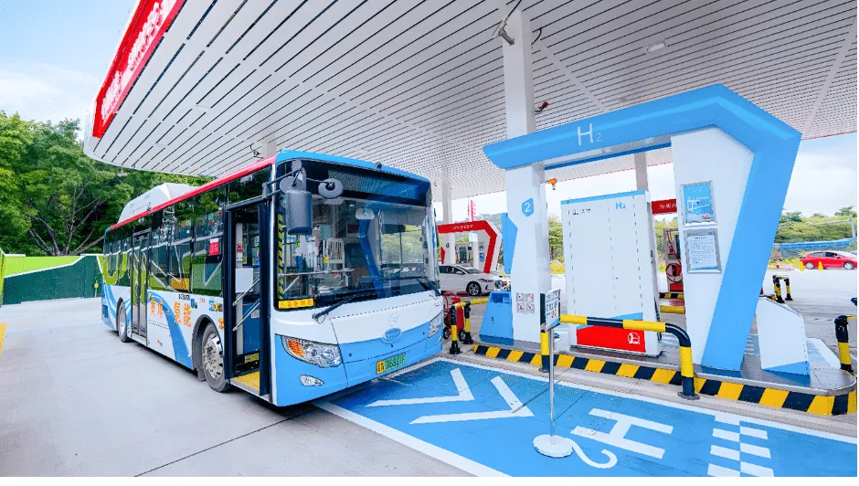 Blue and white city bus at a hydrogen fueling station with an H2 pump nearby under a canopy, marking on the ground.