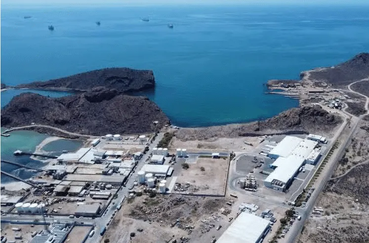 Aerial view of a coastal industrial park with warehouses along a shoreline and rocky hills, blue sea beyond.