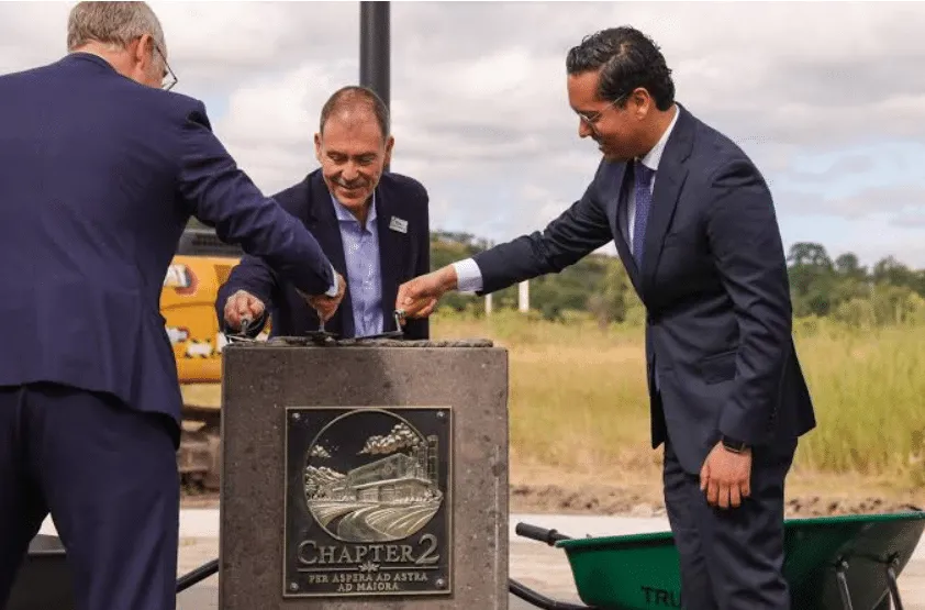Three men in suits participate in a ceremonial lever-touching at an outdoor groundbreaking, around a stone pedestal with a 'Chapter 2' plaque.