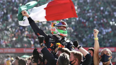 Race car driver wearing a helmet lifts a Mexican flag while being carried by cheering fans in a stadium crowd.