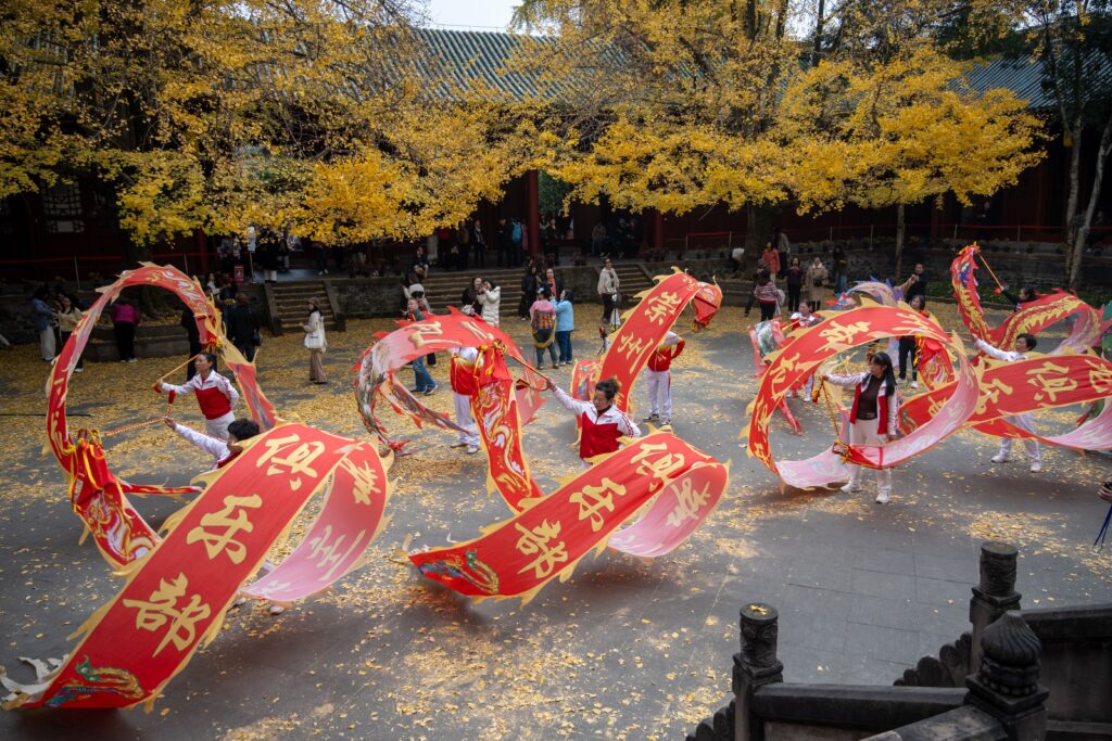 Credits_ Kaca Pic - Petróleo y Energía Dancers in red and white perform with large red banners bearing gold Chinese characters in a courtyard, autumn leaves all around.