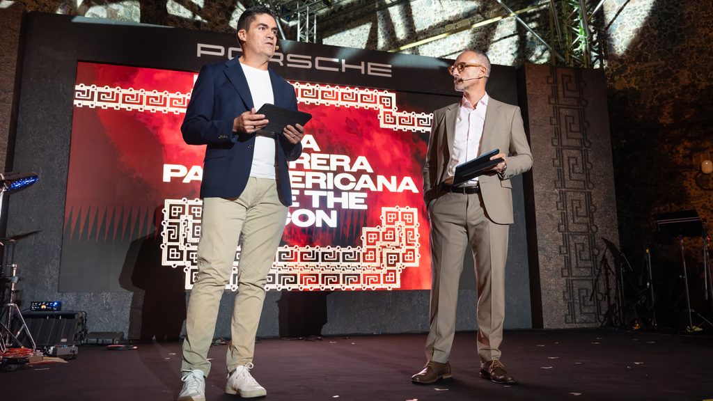 DSC09895-2 - Petróleo y Energía Two men in suits stand on a stage with a red Porsche backdrop, one holding a tablet and the other a folder while speaking into a headset mic.",