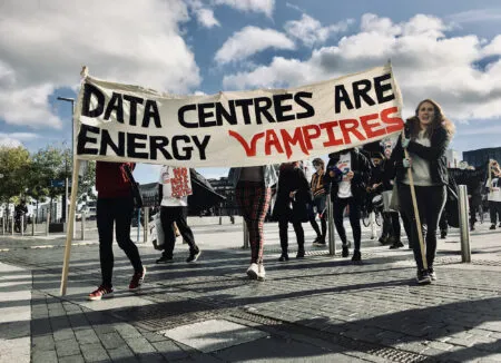 Group of protesters marching outdoors holding a banner that reads: 'DATA CENTRES ARE ENERGY VAMPIRES' under a blue sky with clouds.