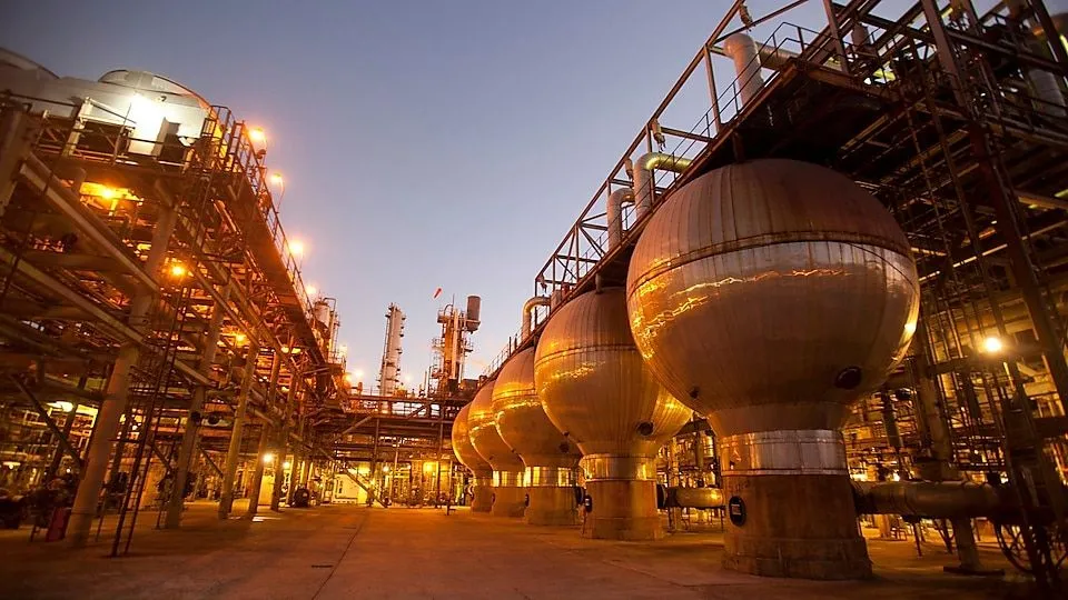 Night view of an industrial refinery with large spherical storage tanks and extensive metal scaffolding, lit by orange lights at dusk.