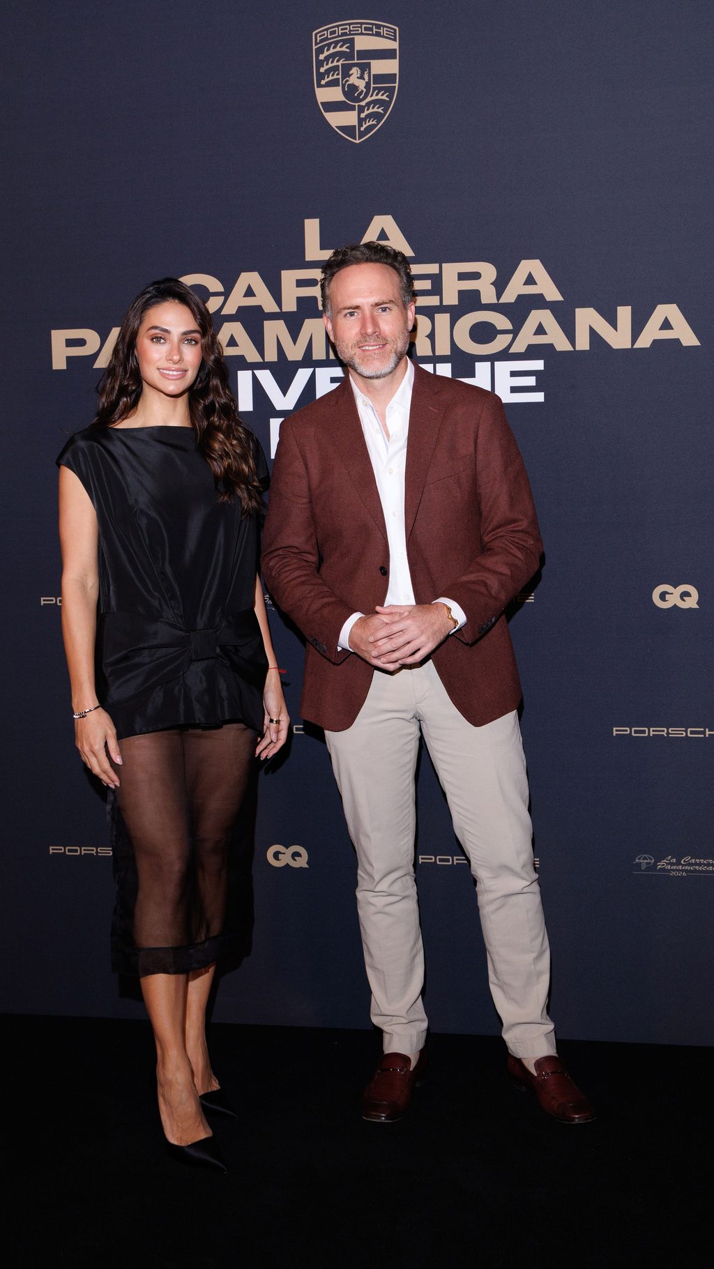 _ETF0704 - Petróleo y Energía Two well-dressed individuals pose on a dark backdrop featuring Porsche branding and event text, a man in a brown blazer and light trousers and a woman in a black dress.