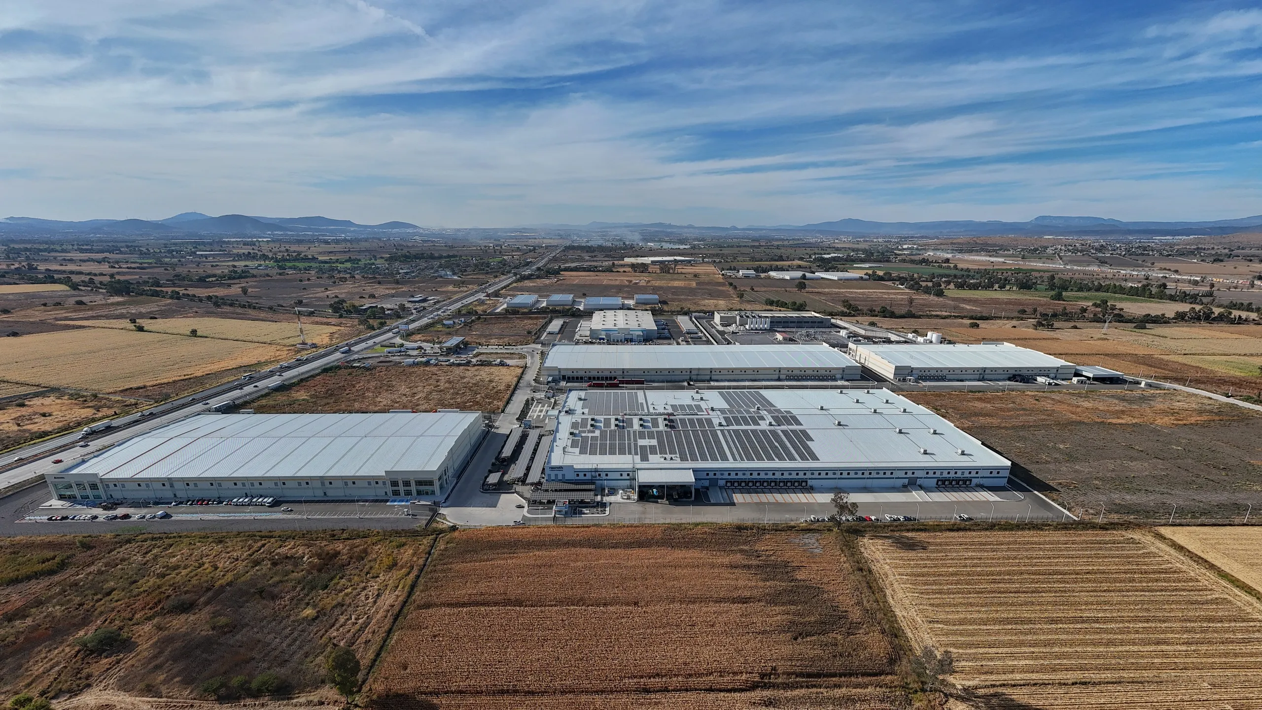 Aerial view of a large industrial complex with white-roofed warehouses and solar panels, surrounded by fields and distant hills under a blue sky.