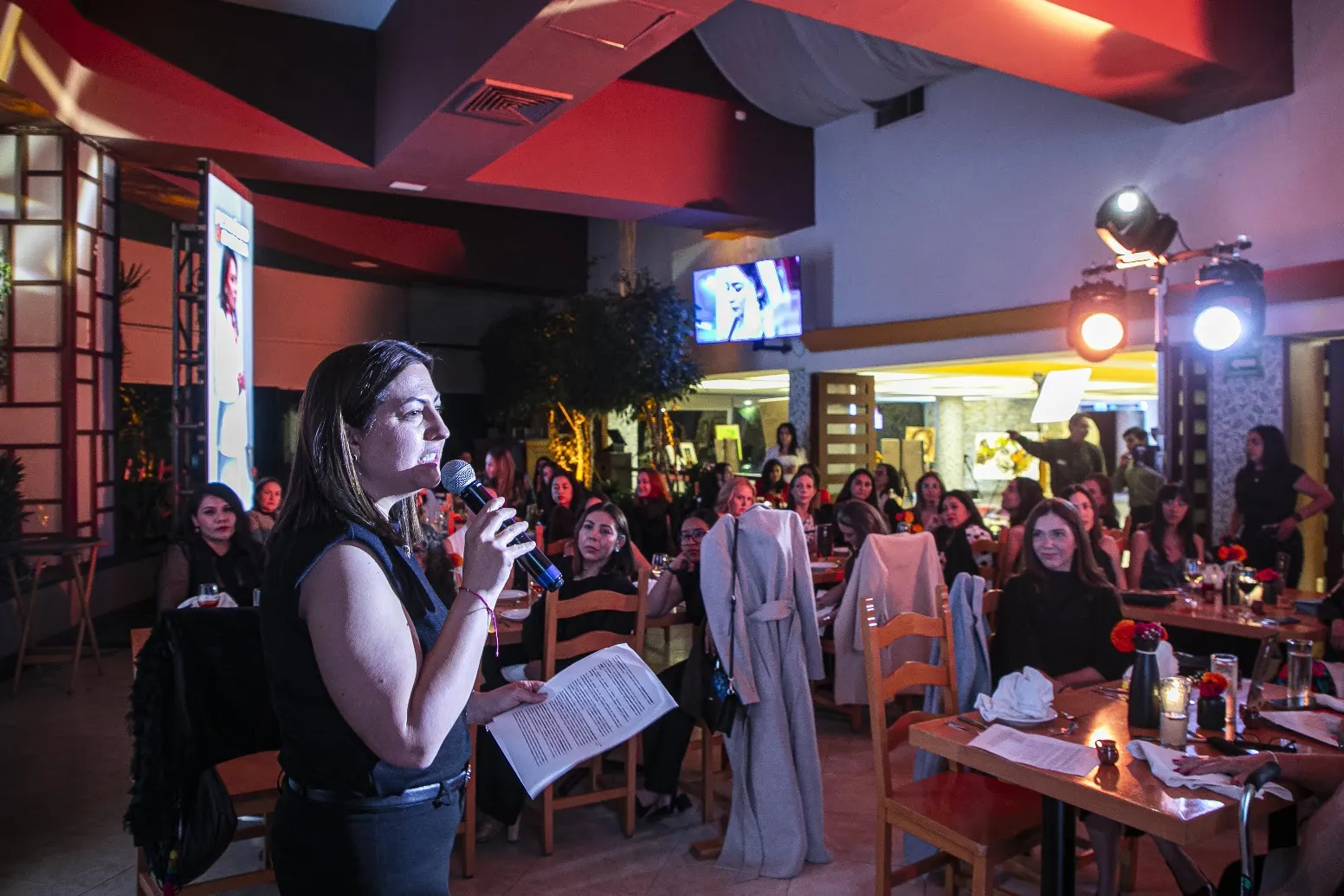 Woman speaker holds a microphone and notes while addressing a dining audience at a restaurant event under colorful lighting.