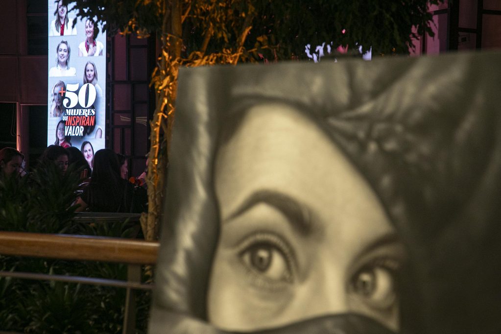 Foto-48 - Petróleo y Energía Blurred statue in foreground with an illuminated poster showing multiple women's portraits and the Spanish text '+50 MUJERES QUE INSPIRAN VALOR' on the left.