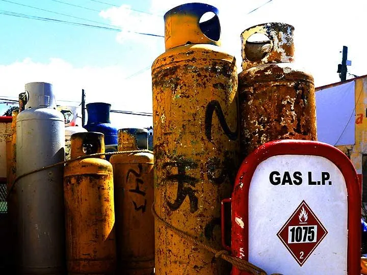 Rusty orange gas cylinders stacked in an outdoor yard, with a red and white Gas L.P. tank labeled 1075 in the foreground.
