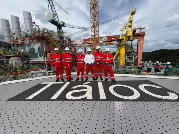 Group of workers in red coveralls and white hard hats pose on a metal platform at an industrial construction site with cranes in the background.