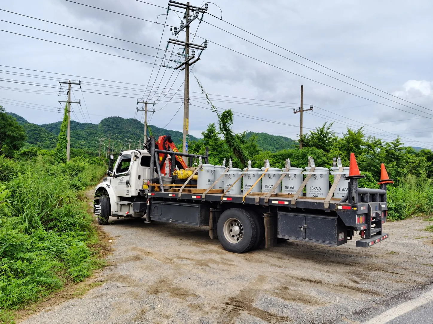 White utility truck with a crane on the flatbed, transporting multiple large gas cylinders along a rural roadside with power lines overhead.