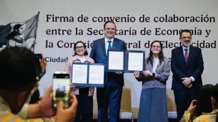 Cuatro ejecutivos en un escenario sosteniendo contratos firmados durante la firma de un convenio de colaboración, mientras la audiencia toma fotos.