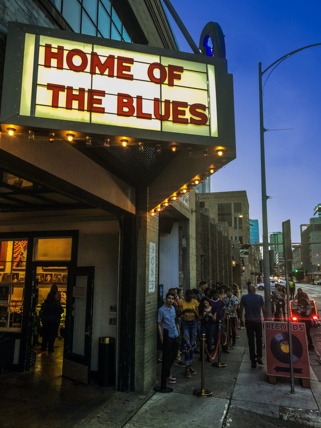 IMG_9047 - Petróleo y Energía Outside a blues venue at dusk, a bright marquee reads'HOME OF THE BLUES' while a line of people waits along the sidewalk.