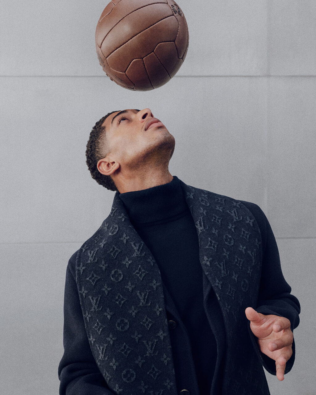 Jude_Bellingham_Louis_Vuitton_Real_Madrid - Petróleo y Energía Man in a dark turtleneck and LV-patterned coat looks up at a basketball suspended above his head due to gravity or a trick shot, mid-motion moment.