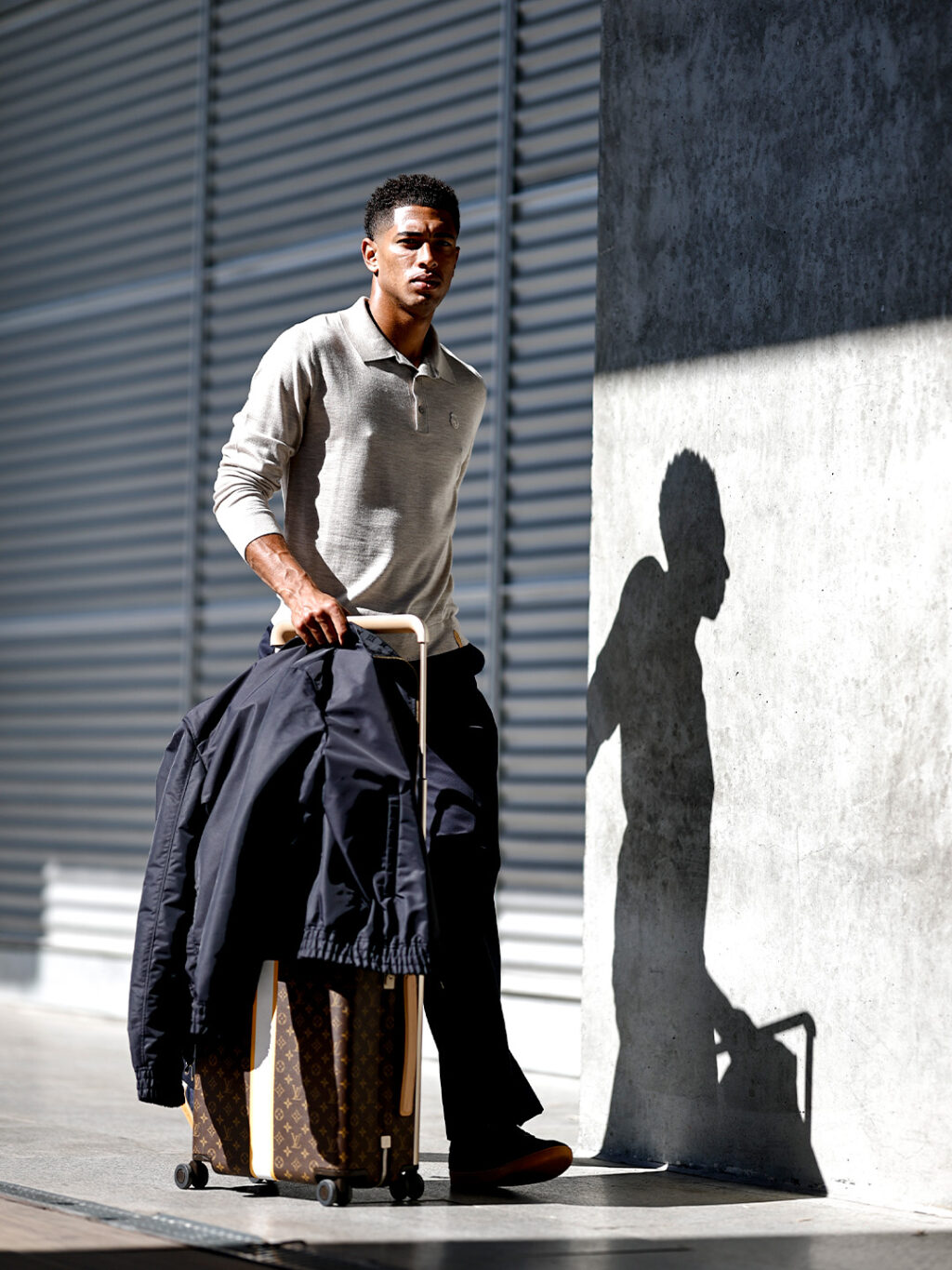 Jude_Bellingham_Louis_Vuitton_Real_Madrid_-_travel_to_Rome_October_2026 - Petróleo y Energía A young man stands on a sidewalk with a rolling suitcase and a navy jacket draped over the handle, beside a concrete wall casting a shadow.
