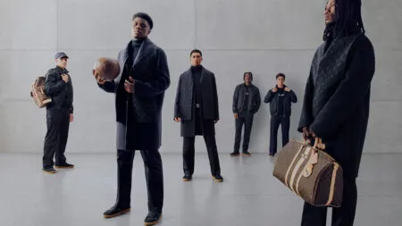 Five men in dark designer outfits pose in a gray studio, each with Louis Vuitton luggage bags arranged around them.