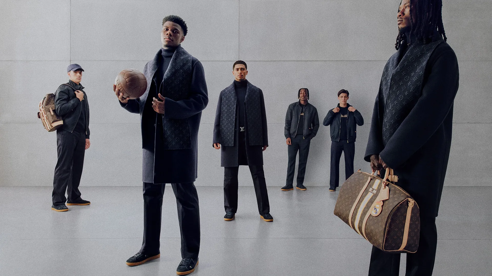 Five men in dark designer outfits pose in a gray studio, each with Louis Vuitton luggage bags arranged around them.