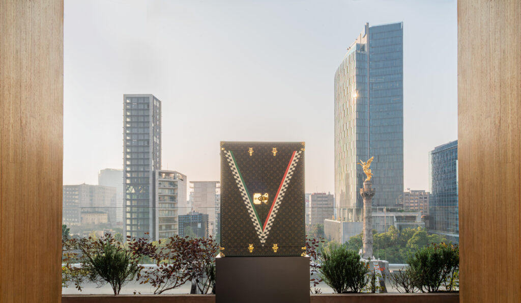 LV-136-2 - Petróleo y Energía Louis Vuitton monogram trunk on a pedestal with a city skyline visible through a large window behind, framed by wood panels .