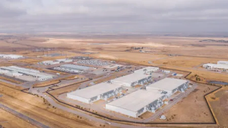 Aerial view of a sprawling industrial park with white warehouses, parking lots, and surrounding brown arid fields under a cloudy sky.