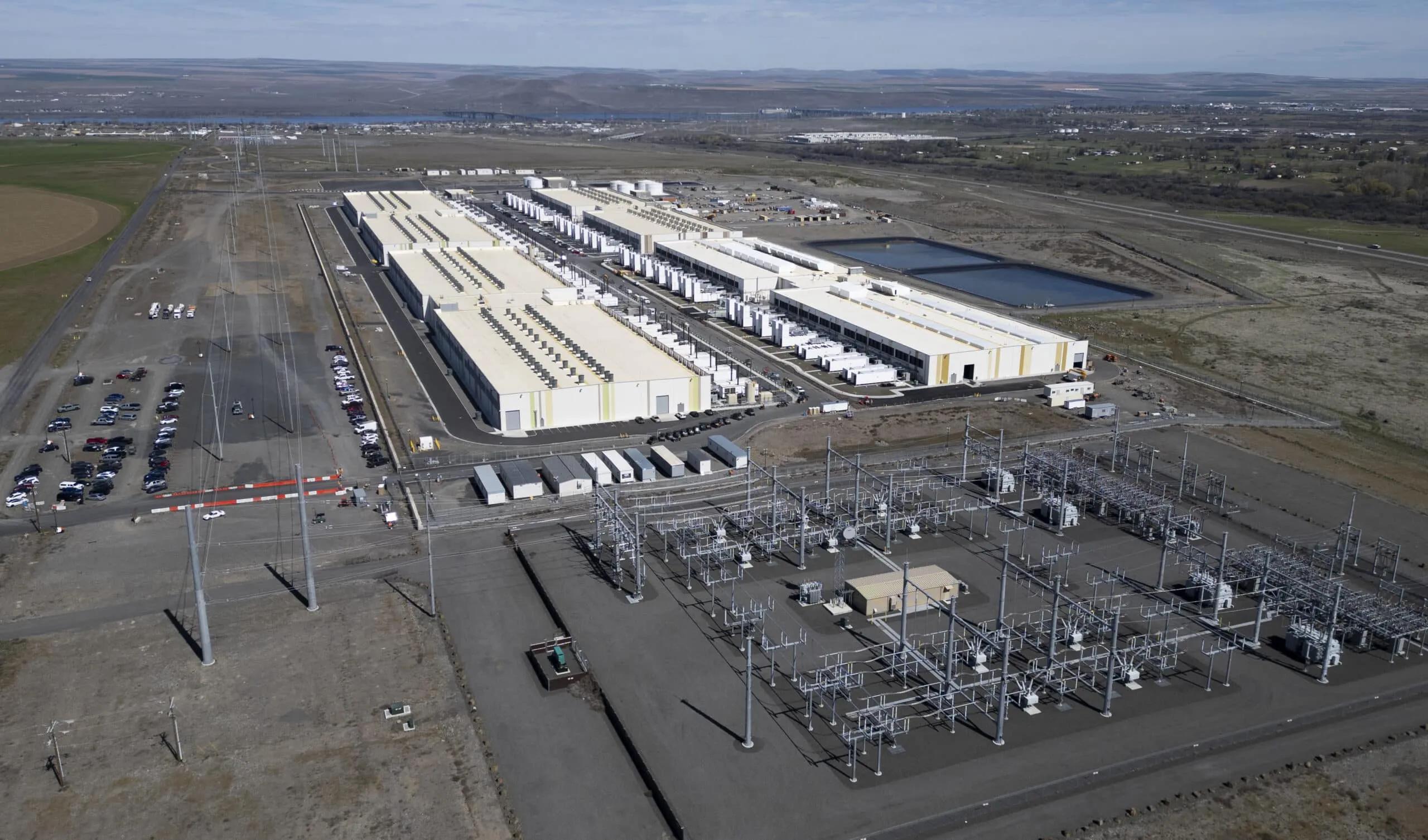 Aerial view of a large industrial complex with long white warehouse buildings, loading bays, parking lots, and power infrastructure in a flat rural landscape.