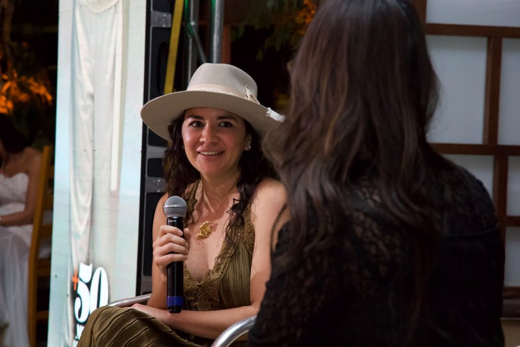 Marielleartista - Petróleo y Energía Smiling woman wearing a beige wide-brim hat holds a microphone and talks to another person at an outdoor event.