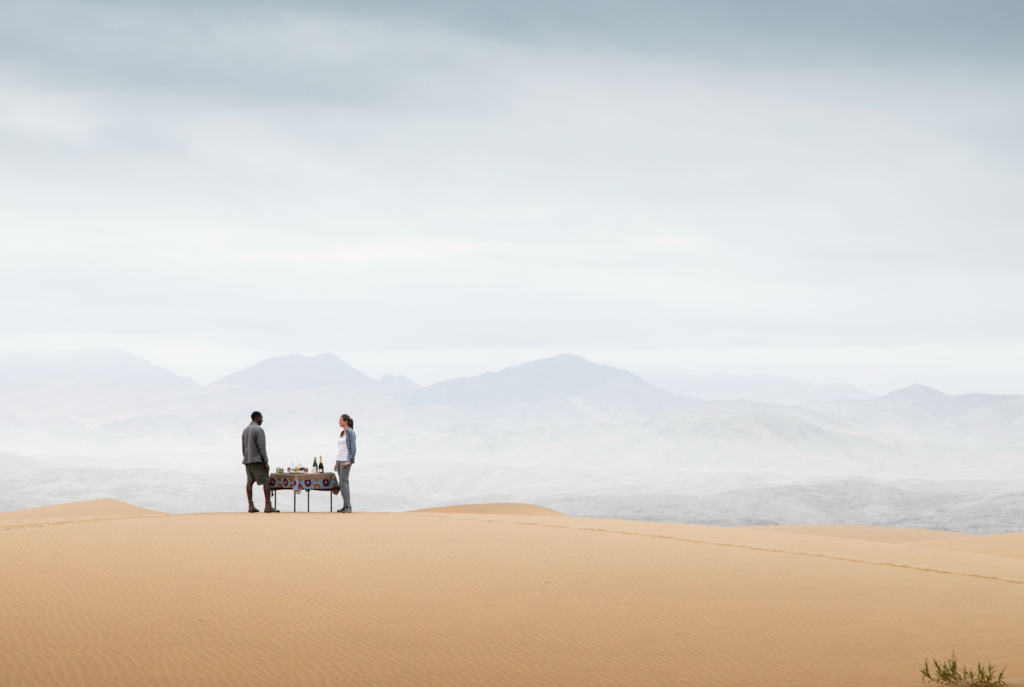 NAMIBIA 2026-10-06 a las 11328 pm - Petróleo y Energía Two people stand on orange desert sand near a small table with drinks, with hazy mountains in the distance under a cloudy sky.
