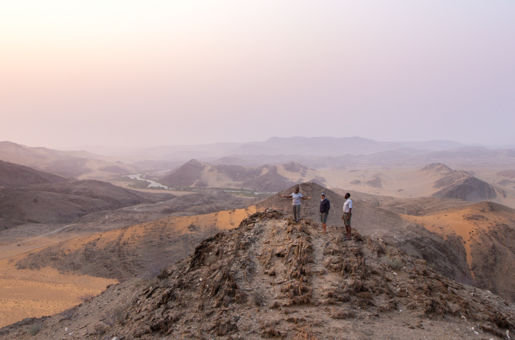 NAMIBIA 2026-10-06 a las 11341 pm - Petróleo y Energía Four people stand on a rocky ridge overlooking a desert valley with distant mountains and a winding river under a pastel pink sky, sunset-like light.