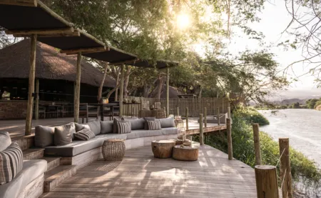 Wooden deck lounge by a river with long beige cushioned seating, woven stools, and thatched-roof huts, sun filtering through trees.