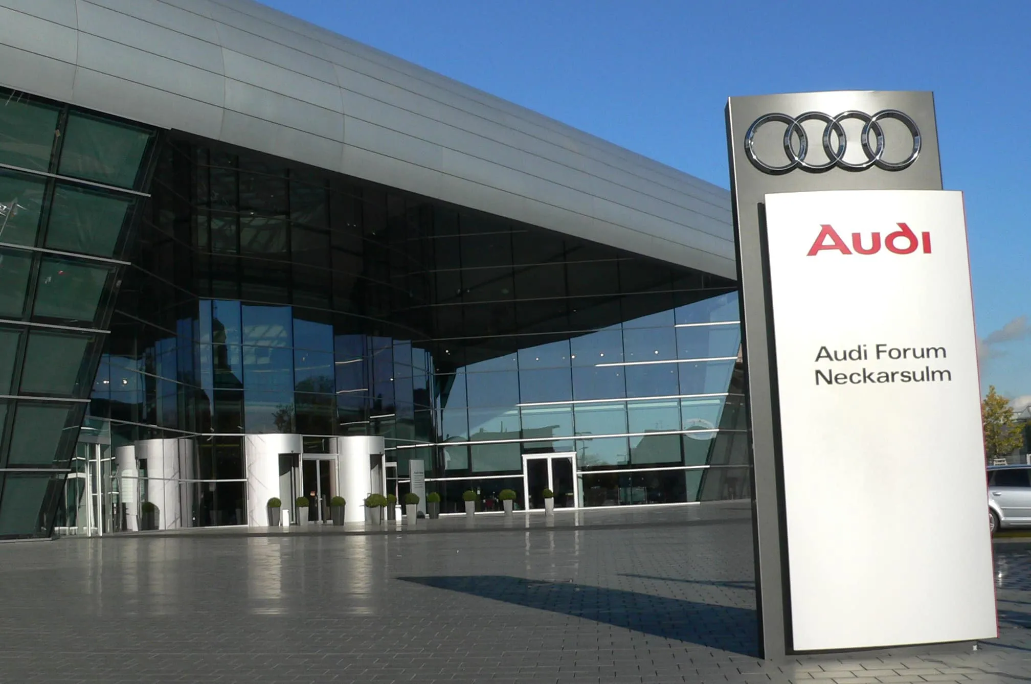 Modern glass Audi Forum Neckarsulm exterior with a tall sign displaying the Audi rings.
