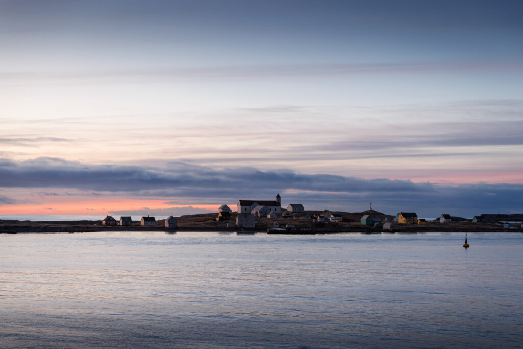 No-2005_CC170125_StPierreMiquelon_Ile aux Marins_©StudioPONANT_MorganeMonneret - Petróleo y Energía Coastal village with colorful houses along the shoreline at sunset, calm water in the foreground and a pastel sky above grafited by light clouds.