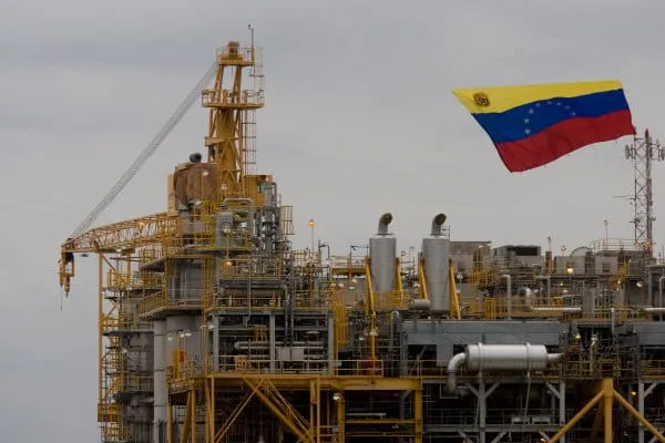 Offshore oil platform with yellow steel framework and a waving Venezuelan flag against a gray sky.