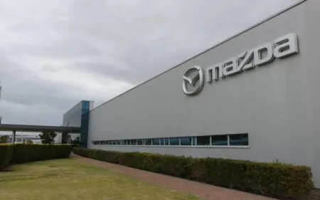 Exterior of a Mazda building with a large Mazda logo on a light gray wall and a hedge-lined walkway