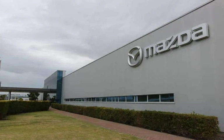 Exterior of a Mazda building with a large Mazda logo on a light gray wall and a hedge-lined walkway