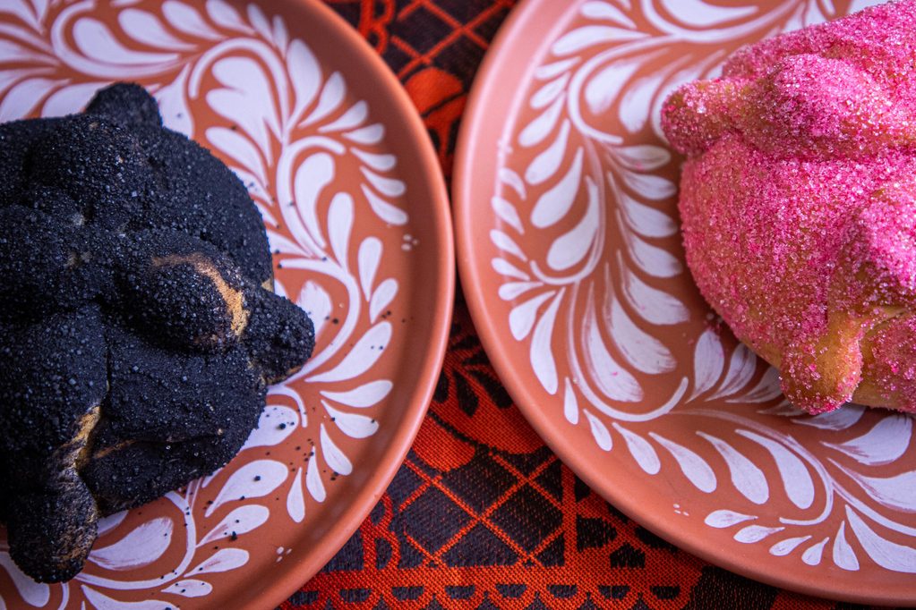 Platillos-6 - Petróleo y Energía Dark sugar-coated cookies on an orange floral plate beside a pink sugar-dusted donut on a decorative orange plate with a red patterned tablecloth behind them.