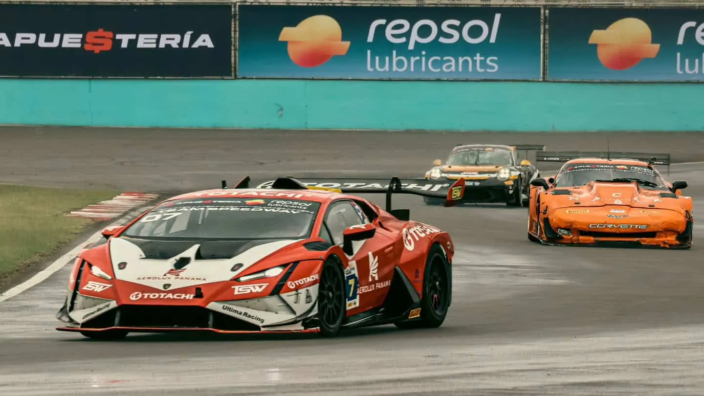 Red Lamborghini race car #7 leads a pack around a bend on a wet track, with an orange Corvette and other cars chasing behind; sponsor banners on the wall.