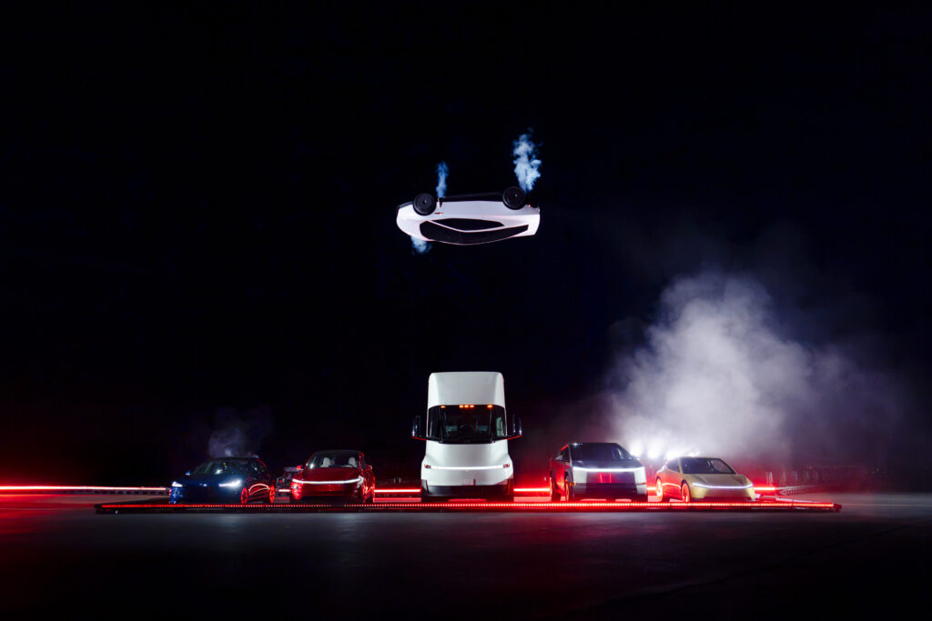 Roadster_58 - Petróleo y Energía White sports car flies midair above a line of vehicles at night with red light trails and smoke in the background, performing a stunt.