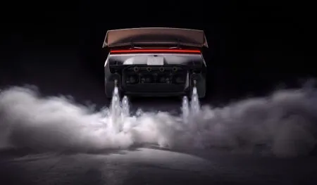 Rear view of a sports car lifting off the ground, large rear spoiler visible, with exhaust plumes and surrounding smoke at night.