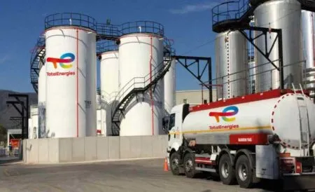 Industrial facility with large white storage tanks bearing TotalEnergies logos and a TotalEnergies tanker truck in the foreground.